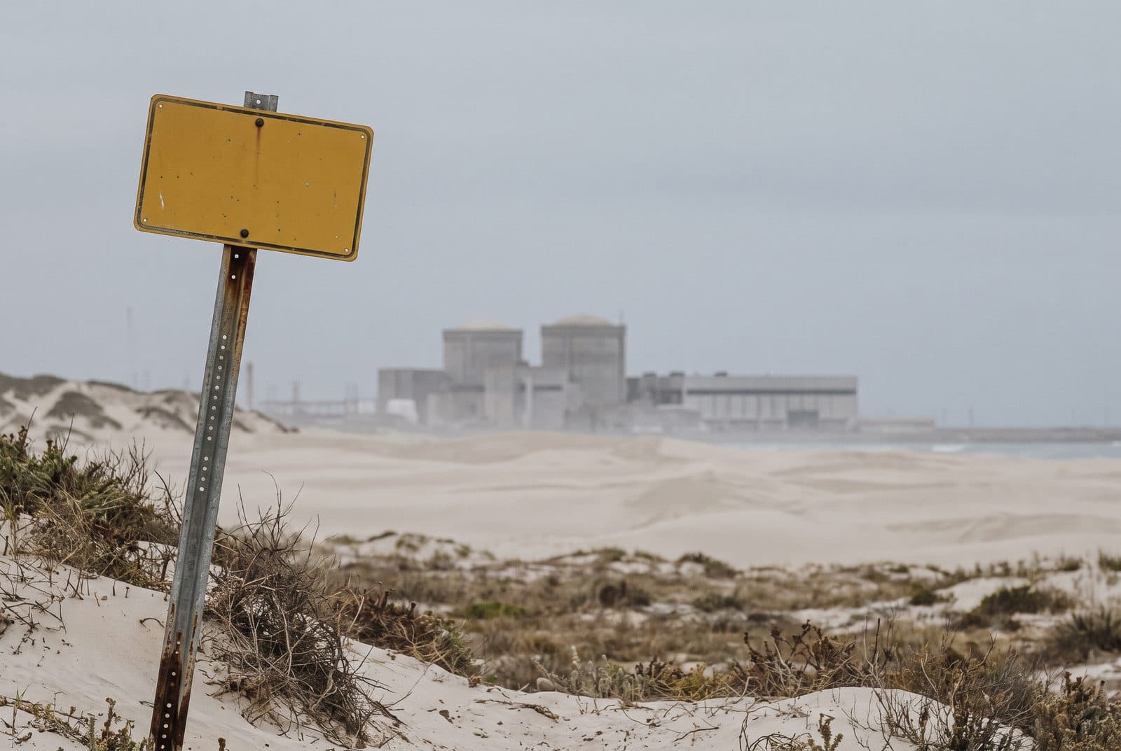 ’n Verweerde geel waarskuwingsbord op ’n staalpaal in die duine, met die kernkragstasie se twee koepels in die dynserige agtergrond.