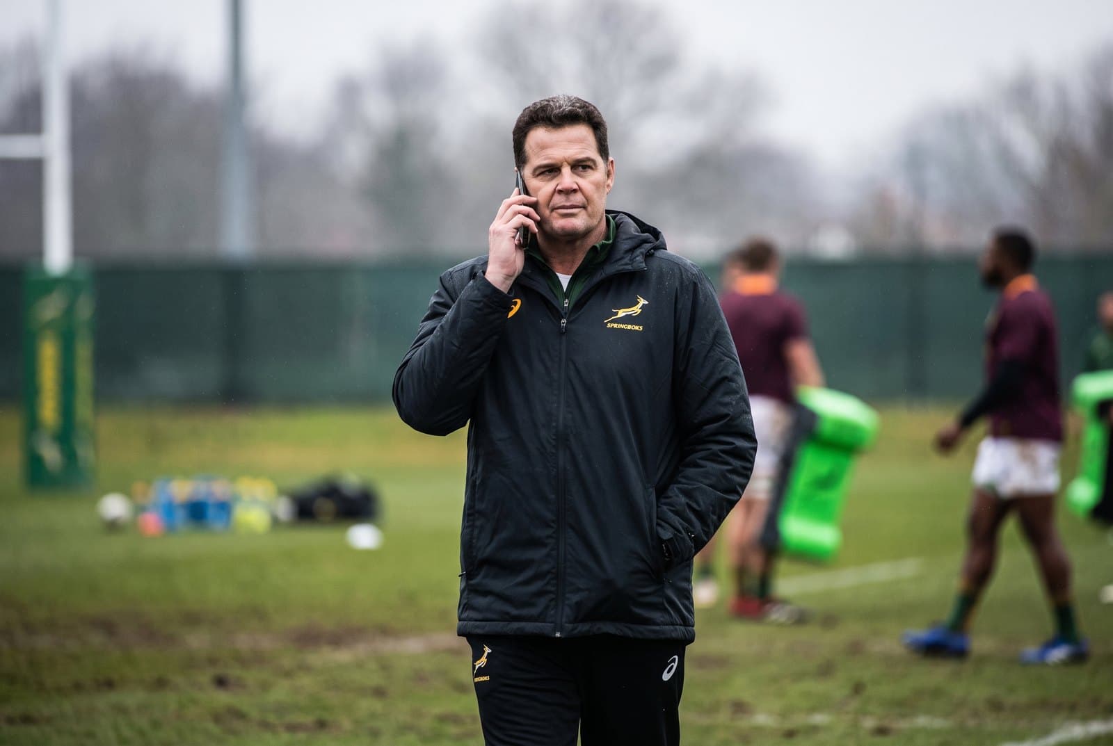'n Rugby-afrigter in Springbok-drag staan op 'n oefenveld met 'n selfoon teen sy oor.